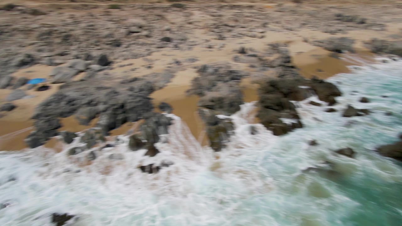 Motion-blurred aerial moment at Buzzards Beach with foamy waves crossing shoreline rocks and sandy beach, with no visible people.