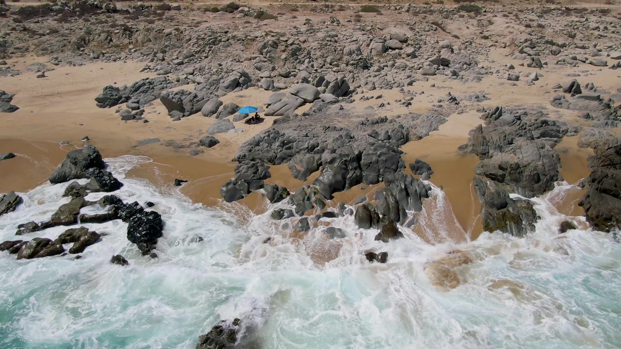 High coastal view at Buzzards Beach showing white foam spreading around dark rocks, dry tan sand beyond, turquoise sea below, and no visible people.