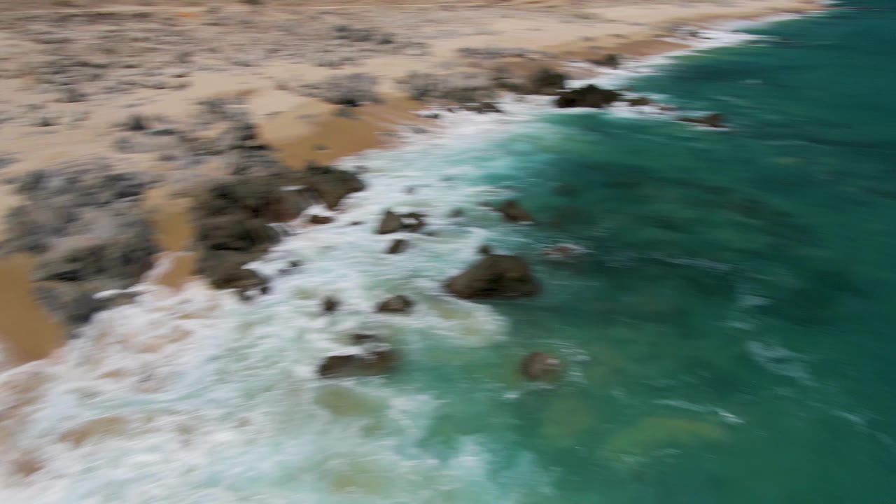 Aerial coastline with surf washing over rocks and deeper green-blue water.