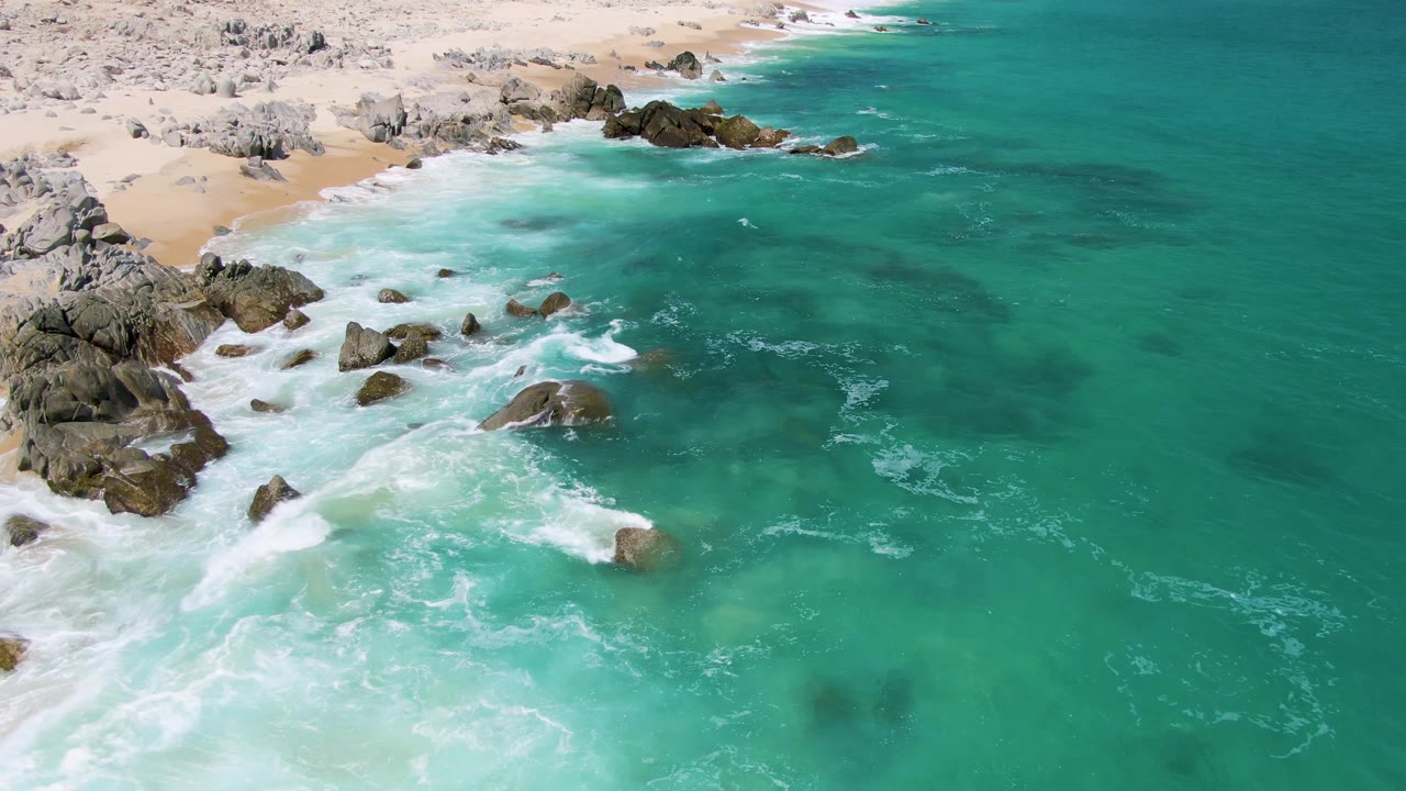 Aerial frame from Buzzards Beach, Baja California Sur, showing turquoise water, white surf around dark rocks, pale sand at the upper left, and no visible people.