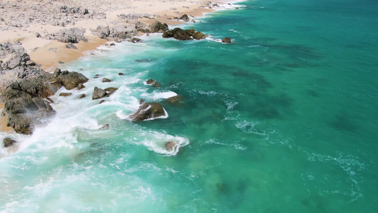 Aerial frame from Buzzards Beach, Baja California Sur, with a larger band of white foam curling around mid-frame rocks and no visible people.