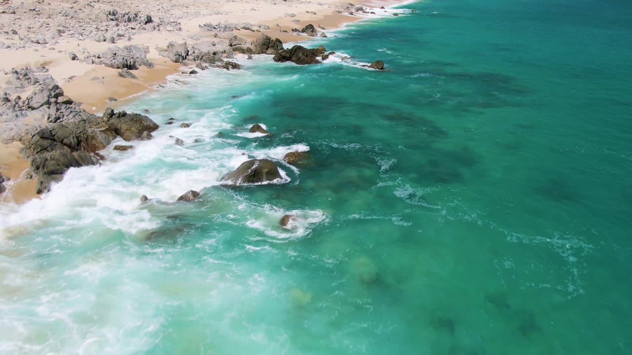 Aerial frame from Buzzards Beach, Baja California Sur, showing bright white surf pushing farther across the rocks, green-blue water to the right, and no visible people.