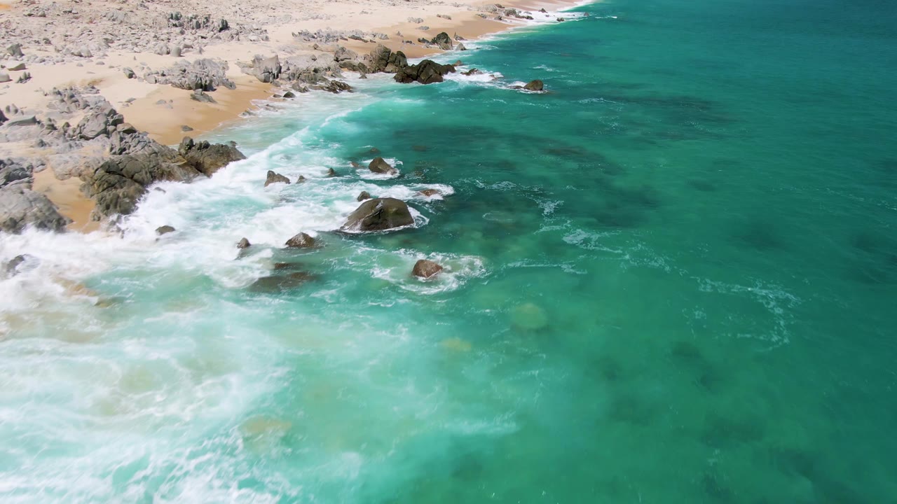 Aerial frame from Buzzards Beach, Baja California Sur, where the rocky shore sits at upper left, foamy water crosses the center, open ocean fills the right, and no visible people appear.