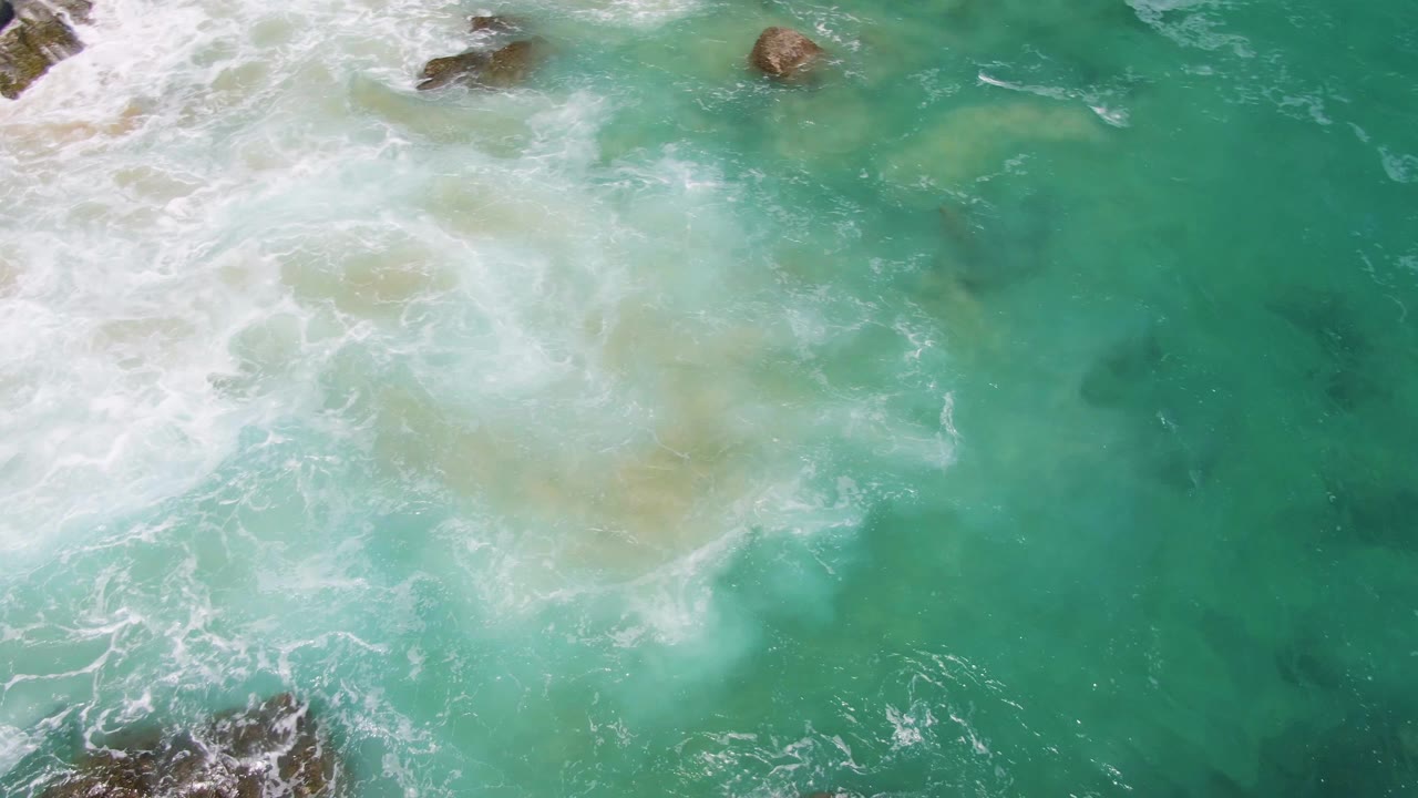 Aerial frame of white surf spreading across turquoise water and rocks at Buzzards Beach, with no visible people.