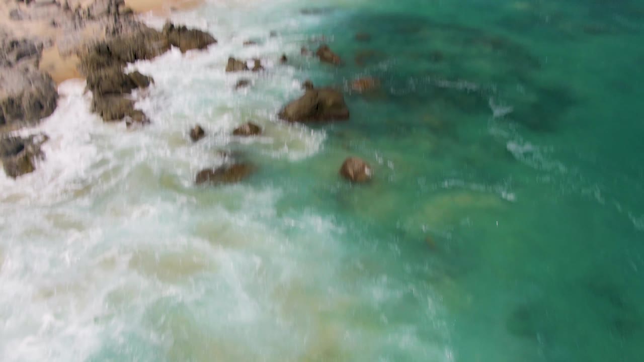 Oblique aerial view of rocks, foamy surf, and clear green-blue water near the Buzzards Beach shoreline, with no visible people.
