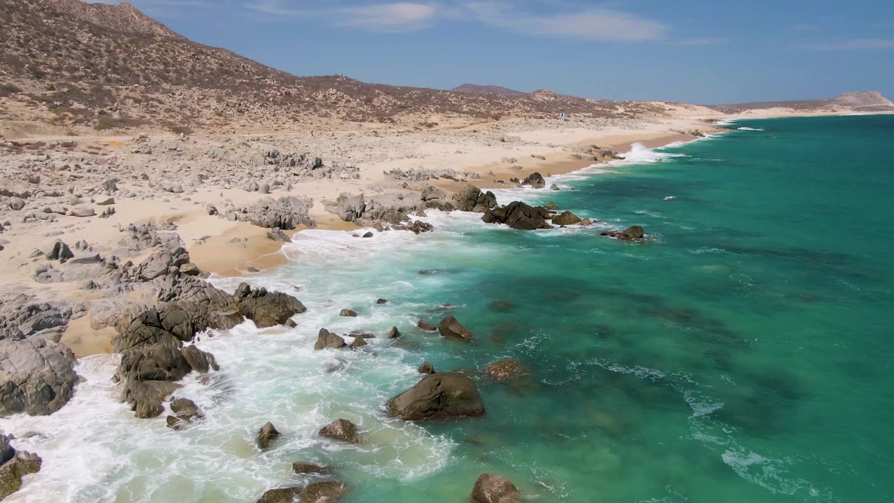 Wide aerial view of Buzzards Beach with a sandy shoreline, dark rocks, turquoise surf, dry hills, blue sky, and no visible people.