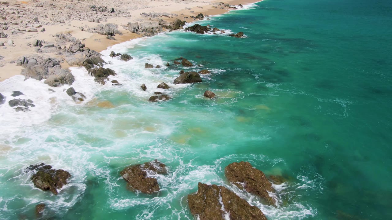Aerial view of Buzzards Beach in Baja California Sur with turquoise water washing around dark rocks, pale sand at the upper edge, and no visible people.
