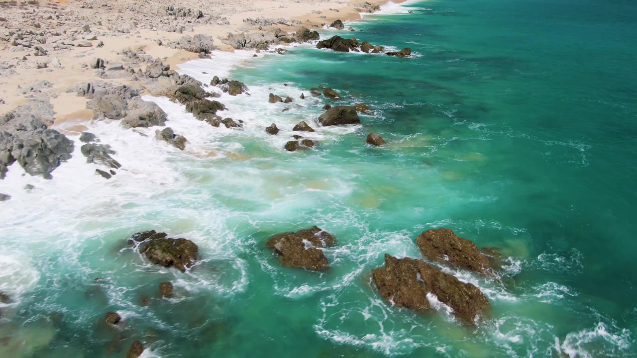 Buzzards Beach shoreline seen from above with several dark rock outcrops in clear turquoise water, breaking white surf near the sand, and no visible people.
