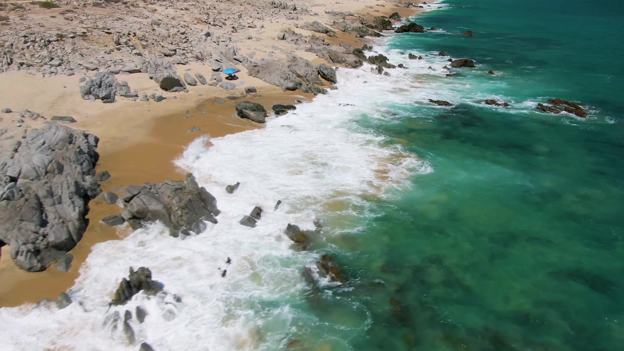 Aerial frame from Buzzards Beach in Baja California Sur showing turquoise water and white surf breaking around dark rocks beside pale sand, with no visible people.