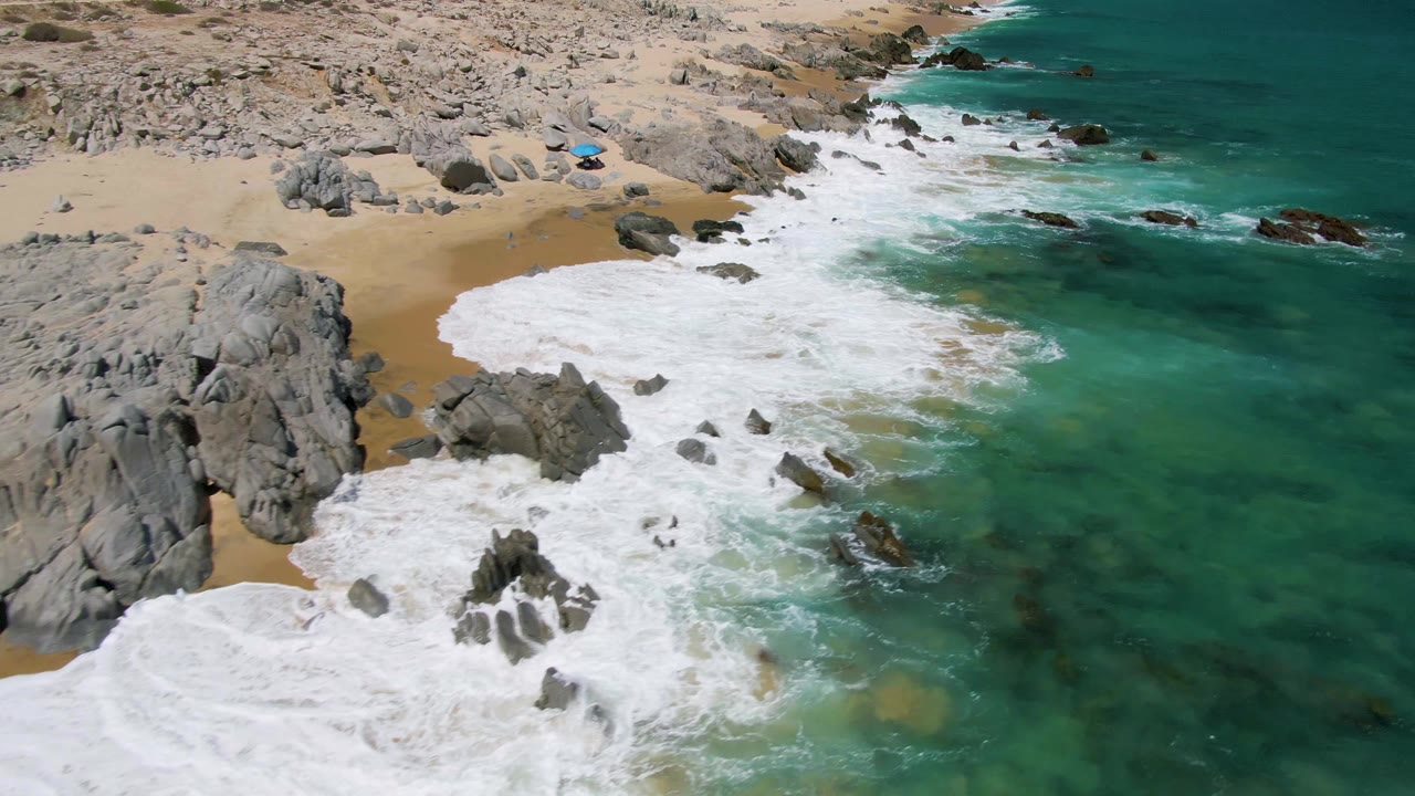 Aerial view over Buzzards Beach with foamy water pushing farther up the sand around clustered rocks and clear turquoise ocean to the right, with no visible people.