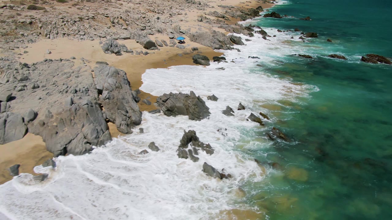 Buzzards Beach shoreline seen from above as a broad sheet of white foam covers the inner rocks while darker underwater patches show through the green-blue water, with no visible people.