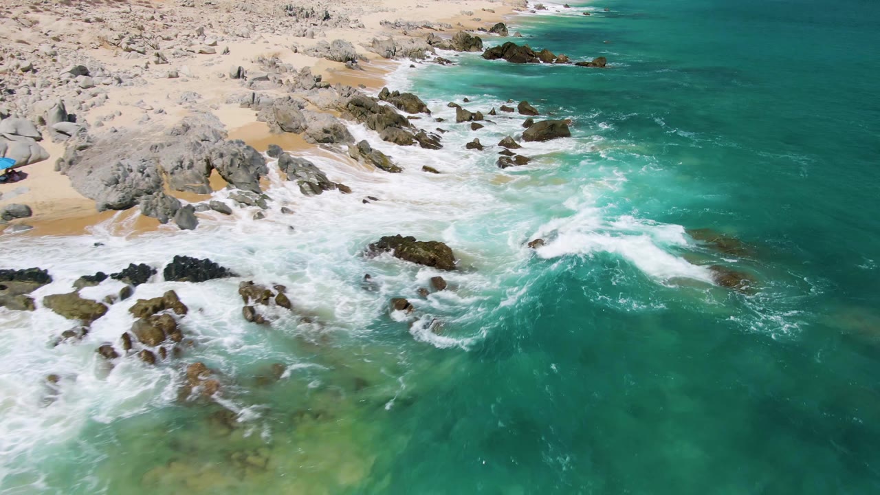 At Buzzards Beach in Baja California Sur, turquoise water breaks into white foam around dark rocks beside tan sand, with pale beach boulders above and no visible people.
