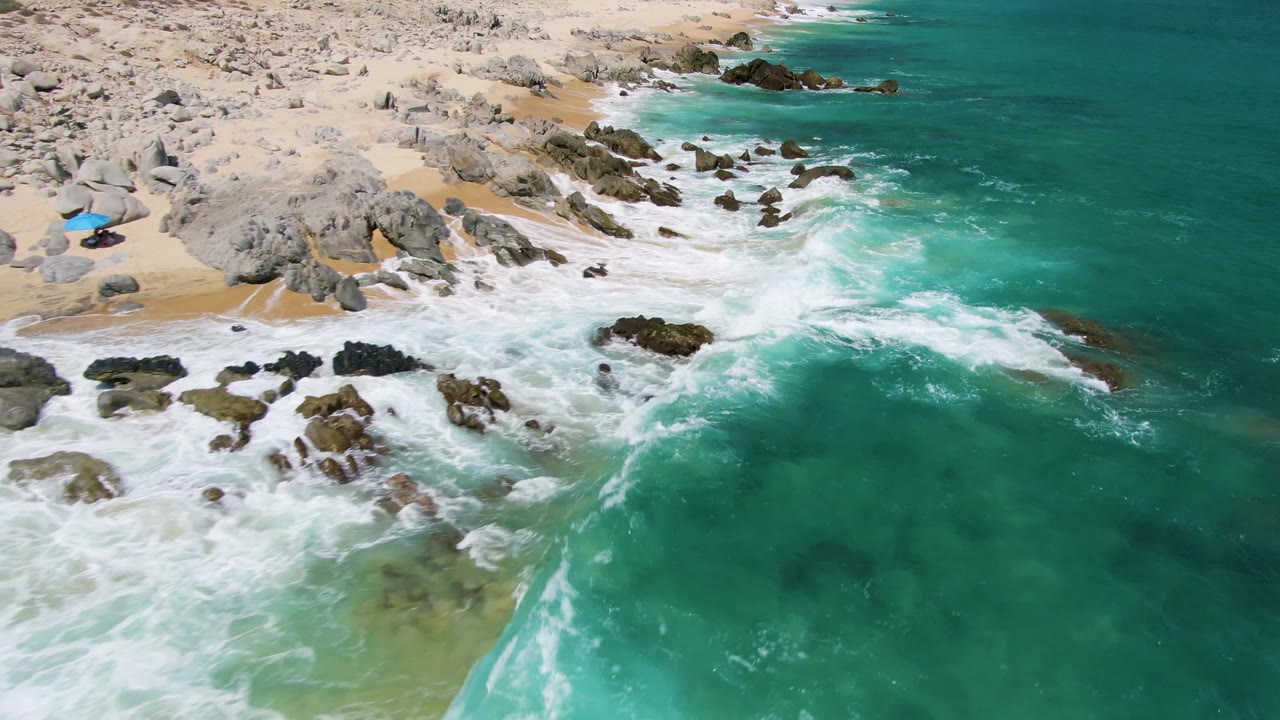 White waves spread across rocks and sand below an aerial coastal view.