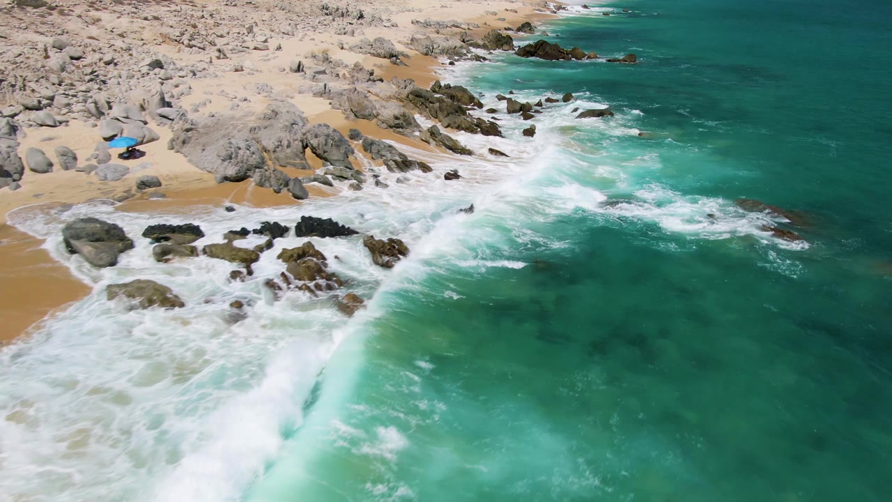 At Buzzards Beach in Baja California Sur, a larger turquoise wave face rises in the foreground as whitewater breaks against rocks and sand near the upper left, with no visible people.