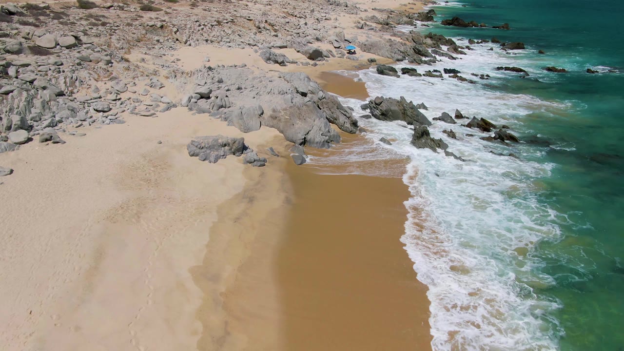 Aerial frame over Buzzards Beach showing a wider strip of wet sand, foamy turquoise surf curling along the right edge, gray rocks above the beach, and no visible people.