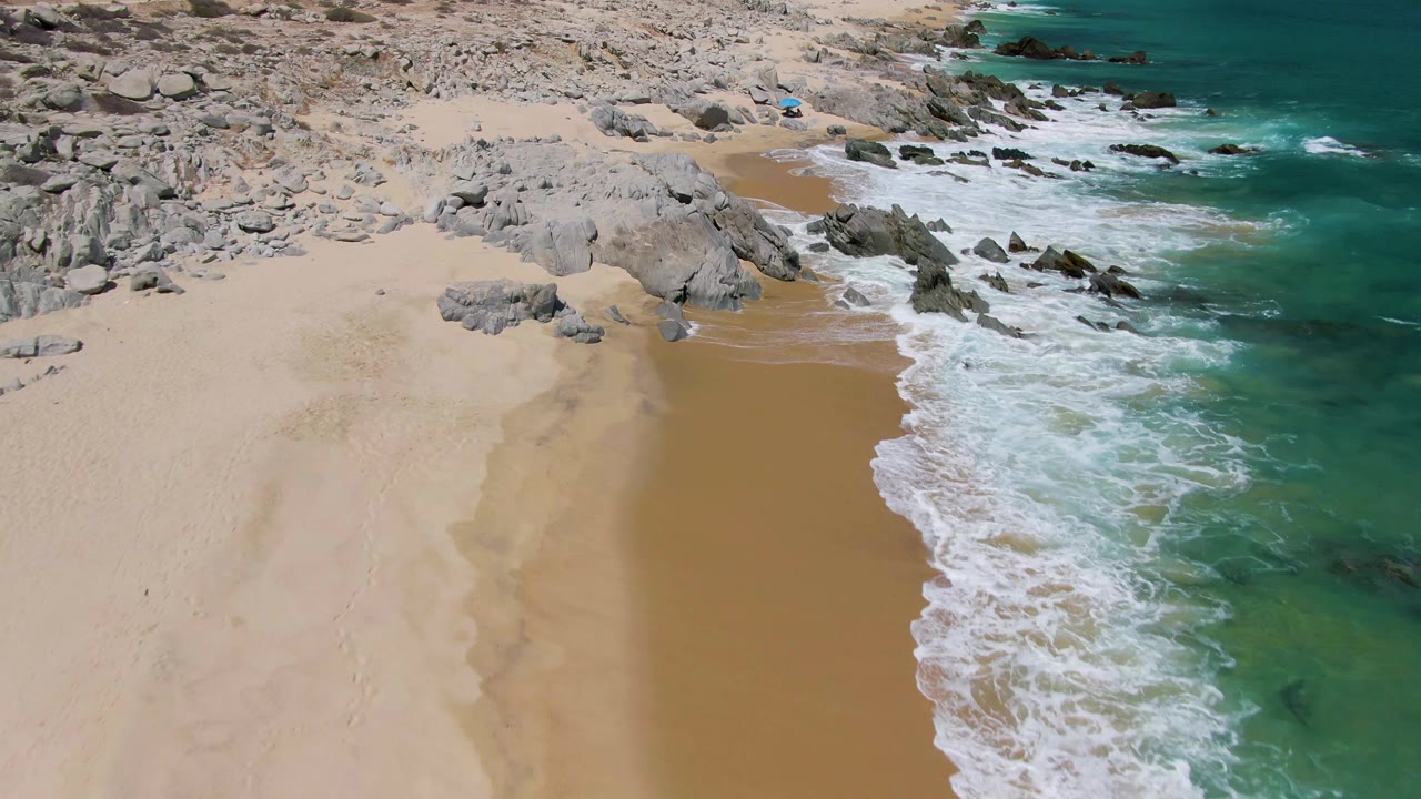 Aerial frame over Buzzards Beach with a broad pale beach on the left, white surf breaking along a darker wet-sand channel, rocky coastline at the top, and no visible people.