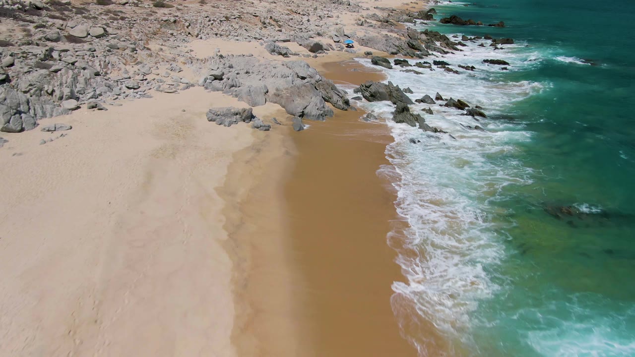 Aerial frame over Buzzards Beach showing the shoreline shifted lower in frame, with foamy waves pushing onto wet sand, large gray rocks at upper left, and no visible people.