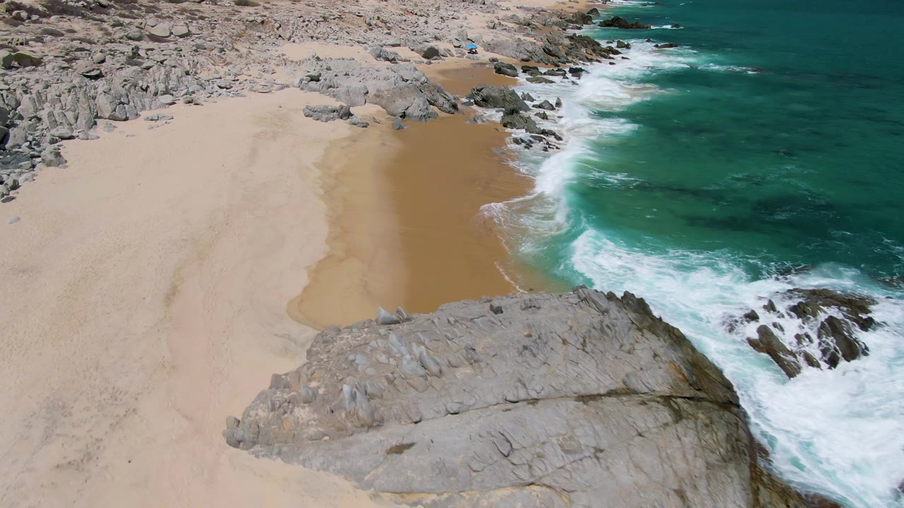Aerial view at Buzzards Beach, Baja California Sur, with turquoise surf touching pale sand beside large gray rock formations and no visible people.