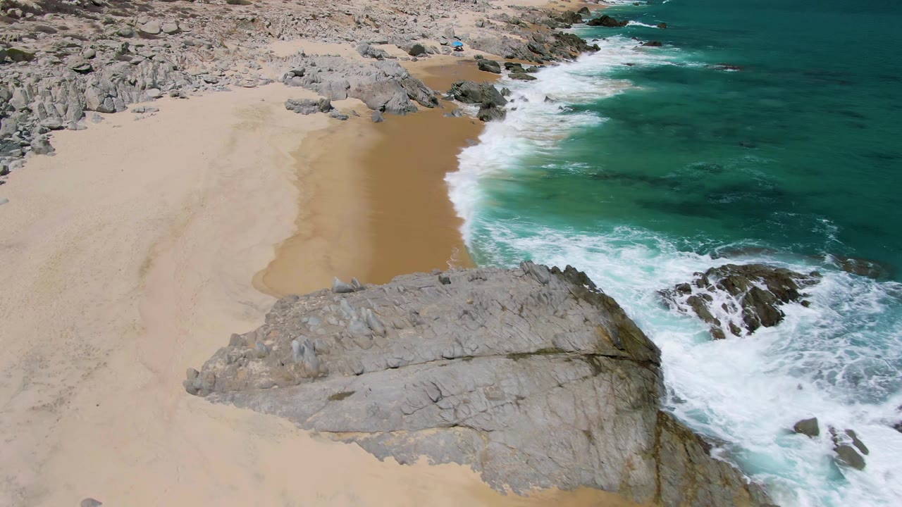 Aerial frame from Buzzards Beach, Baja California Sur, showing a broad gray rock outcrop in the lower frame as white water curls along the sand, with no visible people.