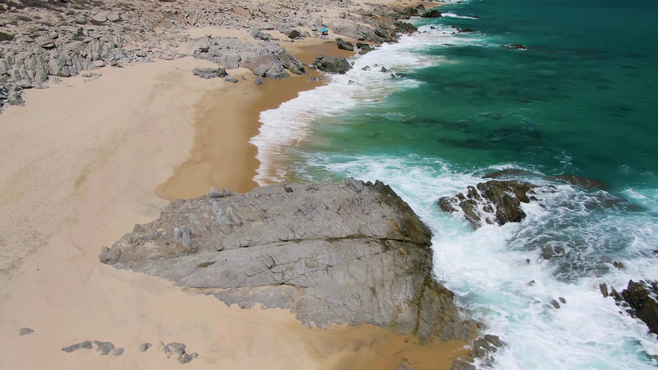 Aerial view at Buzzards Beach, Baja California Sur, with surf wrapping around a central gray rock formation and darker rocks in clear turquoise water, with no visible people.