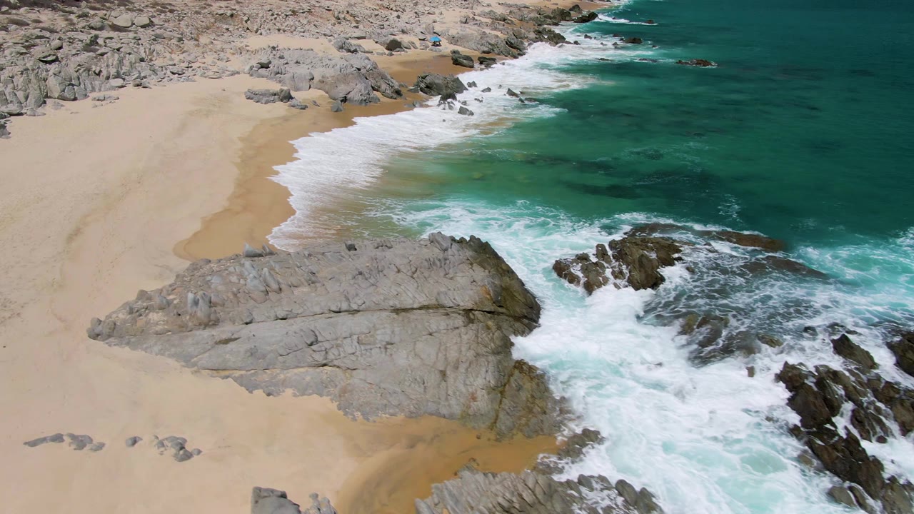 White foam spreads around coastal rocks beside pale sand.