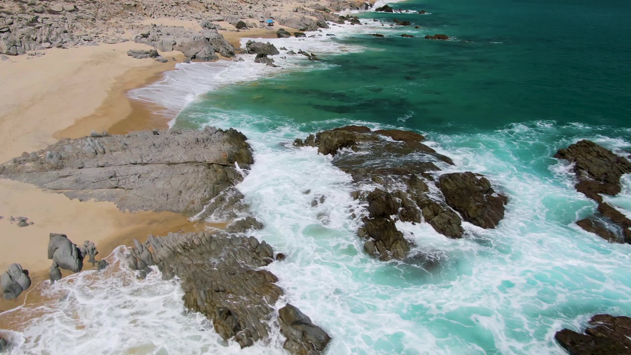 Aerial view of waves breaking over dark rocks beside a sandy beach.