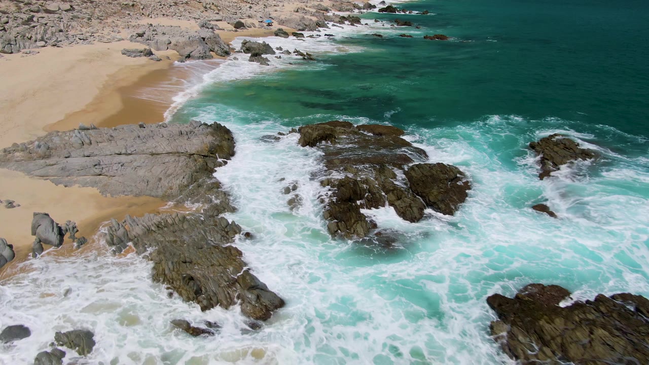 At 0:01, Buzzards Beach surf thickens into white foam across the central rock shelves while the sandy beach remains visible at upper left, with no visible people.