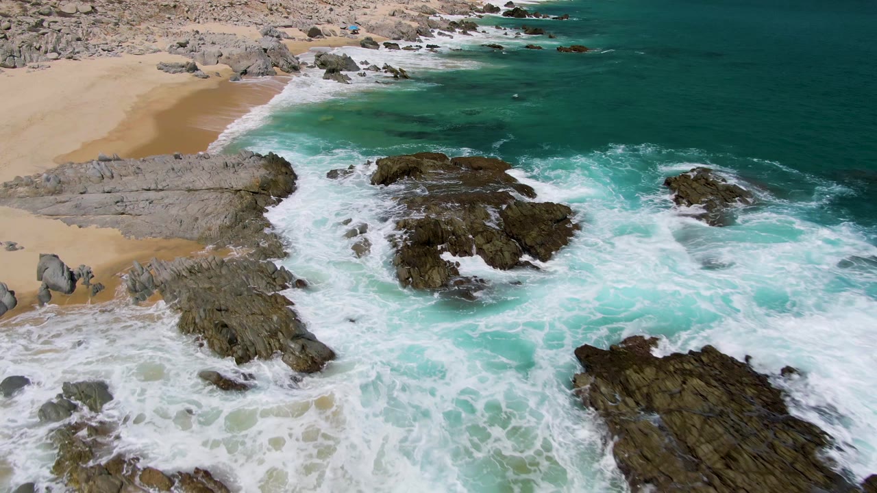 At 0:02, the drone view over Buzzards Beach shows waves curling around several dark rock outcrops with sandy shore and pale rocky ground behind, with no visible people.
