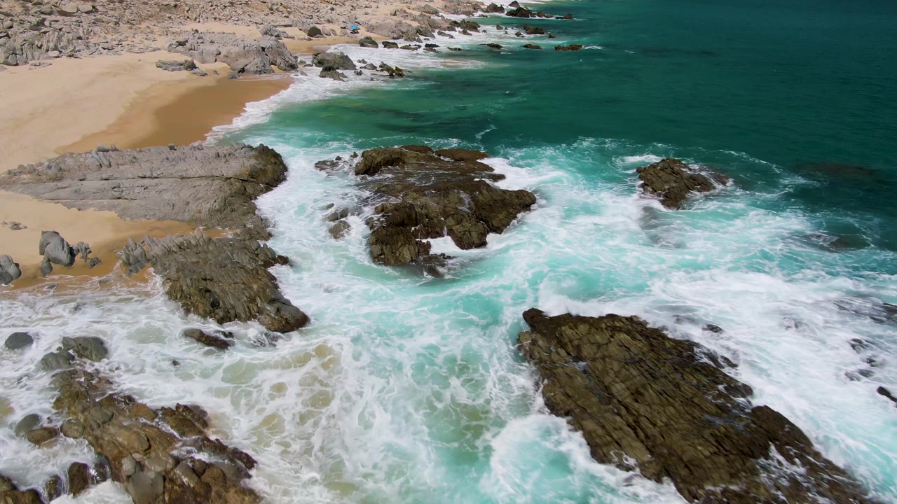 At 0:03, Buzzards Beach surf fills more of the foreground as white foam washes below the central rocks and the sand remains at the upper left, with no visible people.