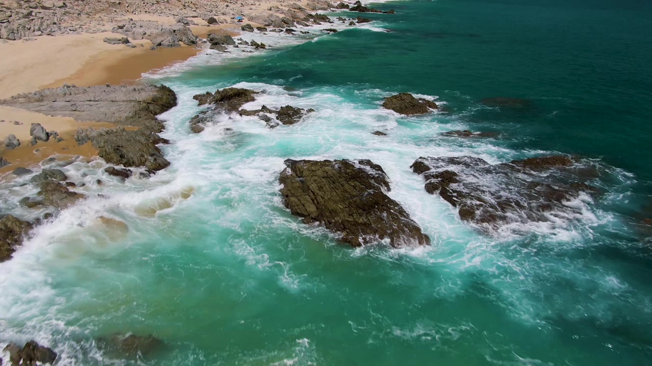Aerial view of turquoise surf breaking around dark rocky outcrops beside the sandy shore at Buzzards Beach, Baja California Sur, with no visible people.