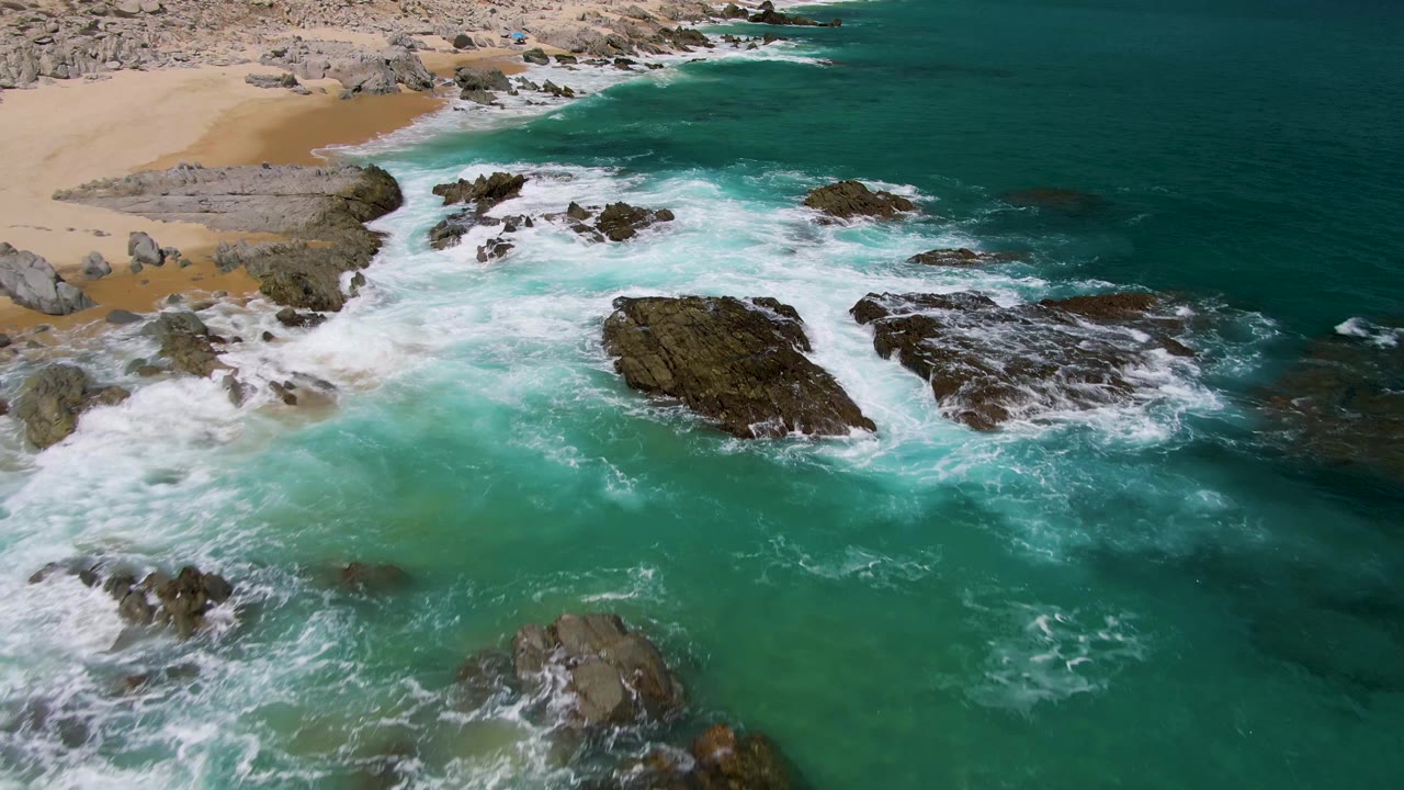 Aerial frame from Buzzards Beach showing whitewater spreading across several dark rocks in shallow turquoise ocean, with the sandy beach at the upper left and no visible people.