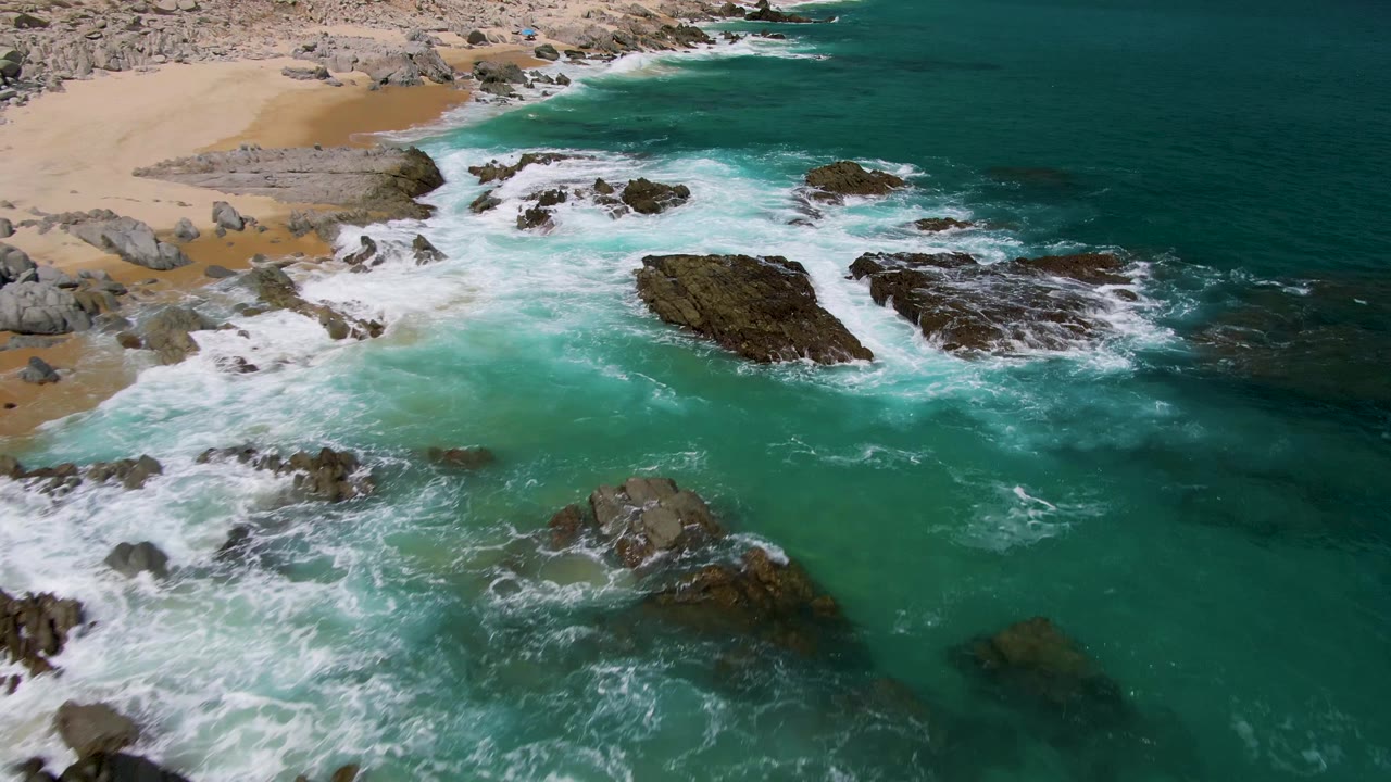 Aerial view at Buzzards Beach with foamy surf curling into a turquoise nearshore pocket, dark rocks offshore, sand and boulders along the top edge, and no visible people.