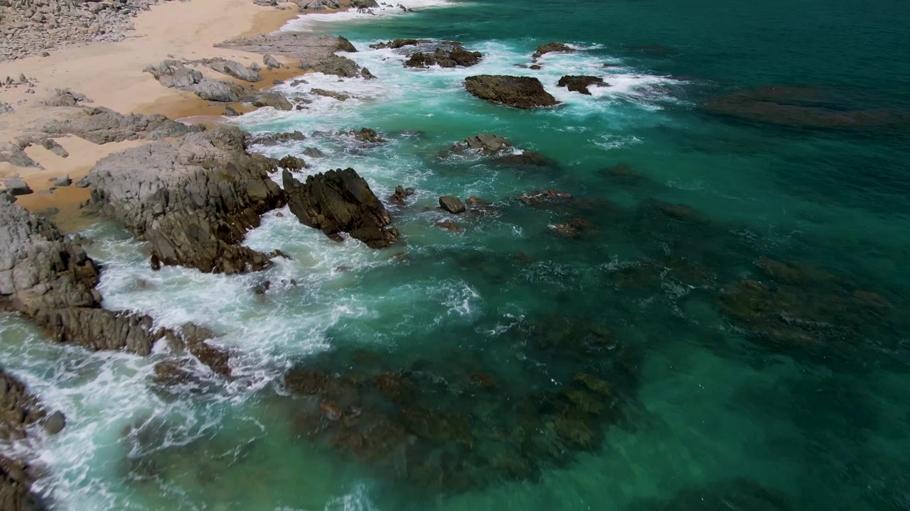 Aerial view of turquoise waves breaking around rocky shoreline and sand