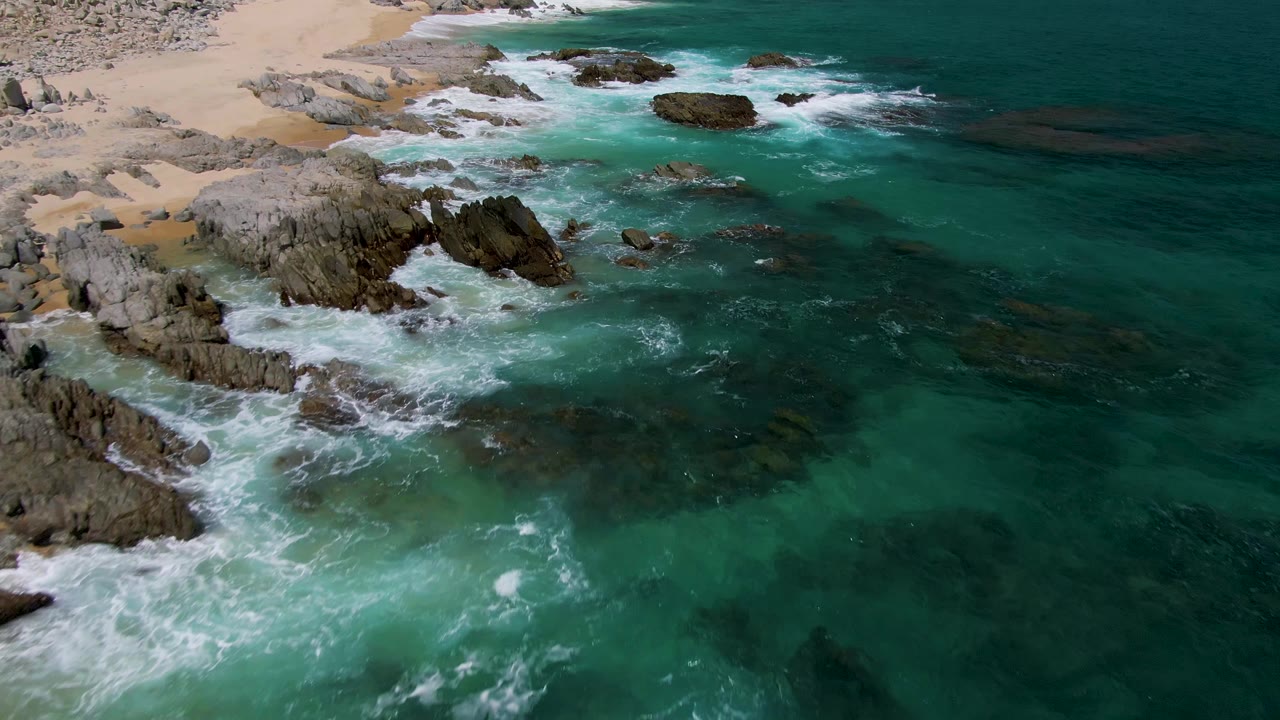 Aerial frame over Buzzards Beach with white foam spreading through shallow turquoise water between rock formations and a strip of sand in the background, with no visible people.