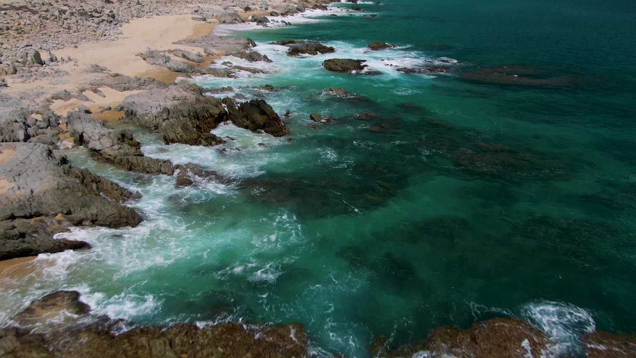 Aerial frame over Buzzards Beach showing open turquoise ocean to the right, rock shelves along the shore, small sandy coves at the upper left, and no visible people.