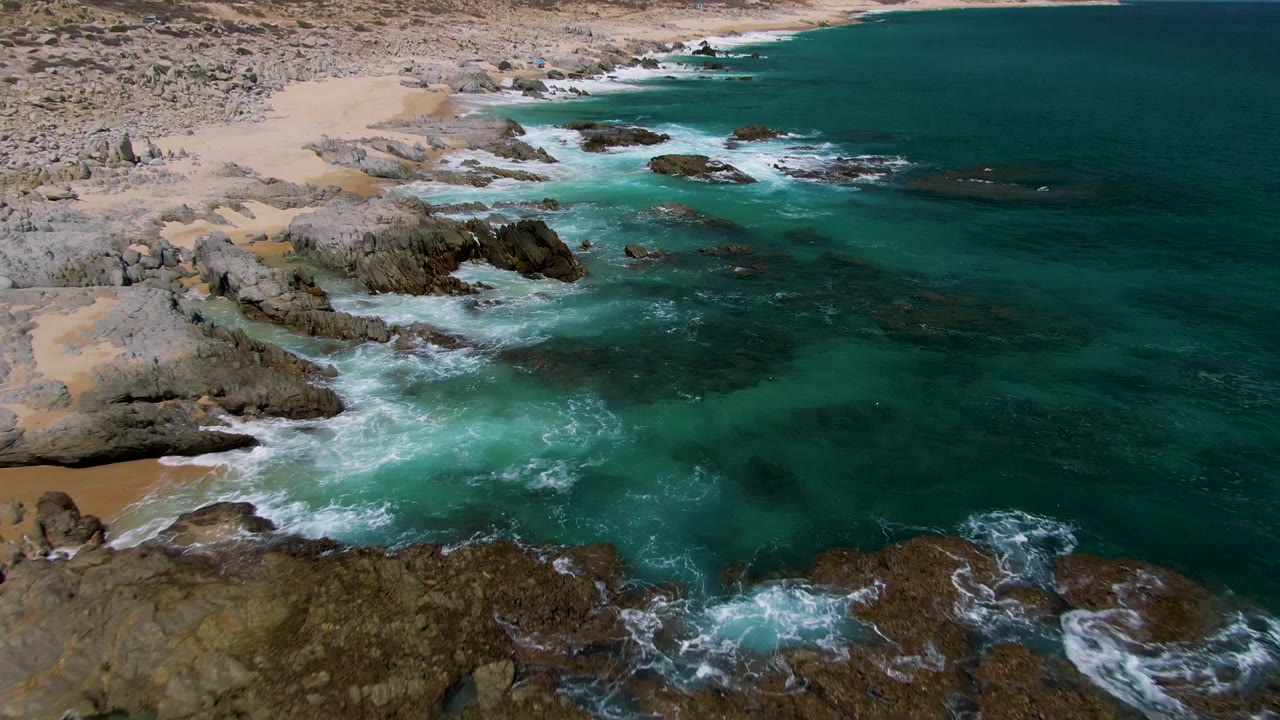 Aerial frame over Buzzards Beach with a broader coastline view, foamy waves over foreground rocks, sandy beach on the left, dark ocean to the right, and no visible people.