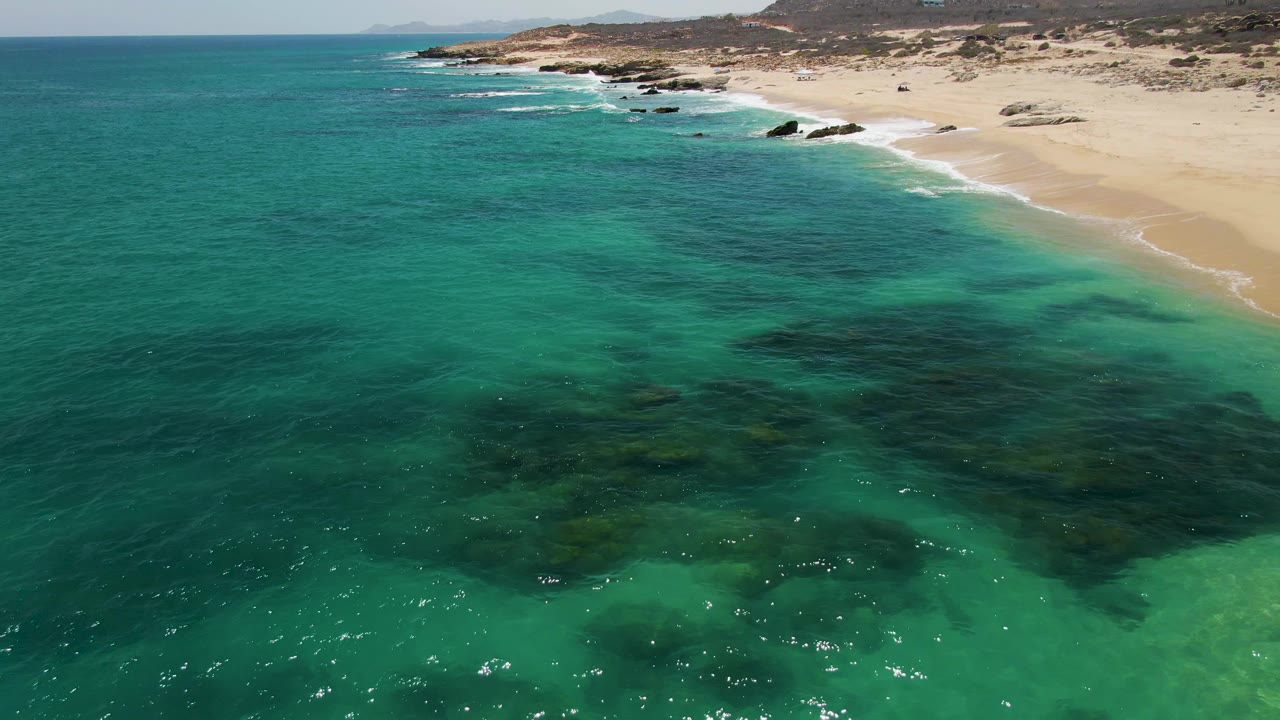 Opening aerial frame over Buzzards Beach, Baja California Sur, with turquoise water in the foreground, pale sand and shoreline rocks beyond, and no visible people.