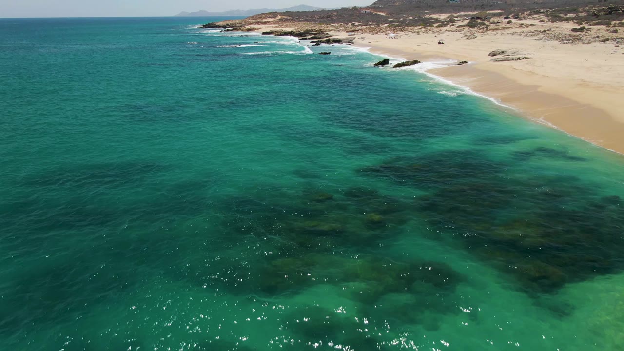 Aerial frame at Buzzards Beach showing sunlit turquoise shallows, dark underwater rock areas, a sandy shore curving across the top, and no visible people.