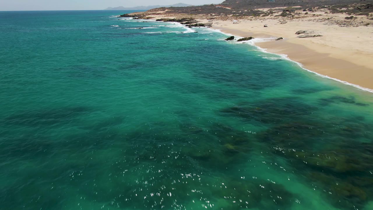 Buzzards Beach aerial view with open turquoise water in front, white foam tracing the sandy shoreline, rocky points along the beach, and no visible people.