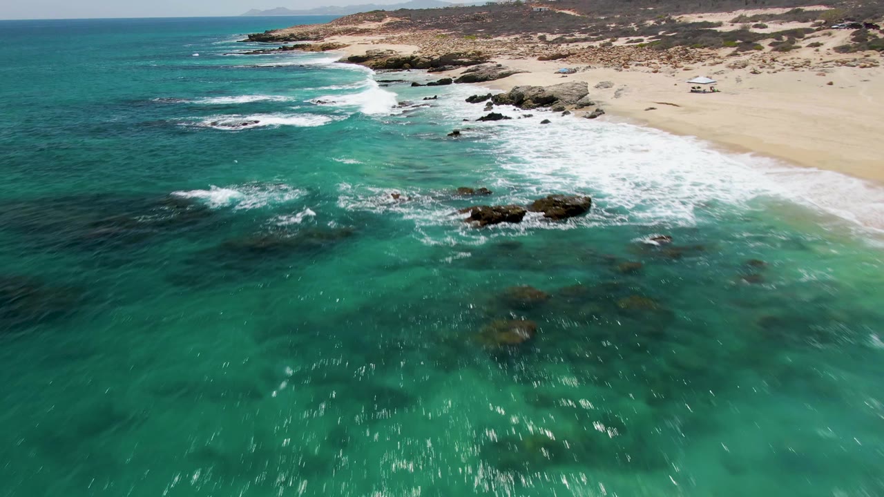 Aerial frame over Buzzards Beach in Baja California Sur, Mexico, with turquoise water in the foreground, white surf on dark rocks, pale sand to the right, distant coastline, and no visible people.