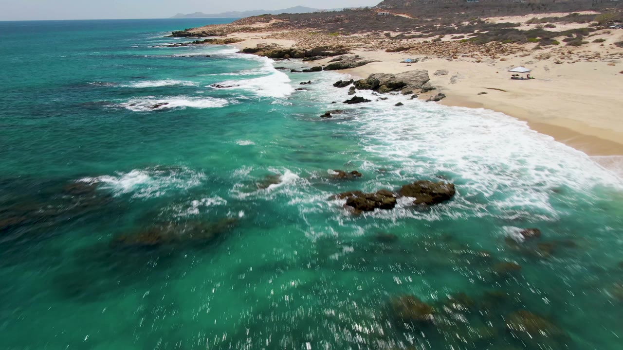 Aerial view at Buzzards Beach in Baja California Sur, Mexico, with broken white water spreading across nearshore rocks, sandy beach on the right, low scrubland beyond, and no visible people.