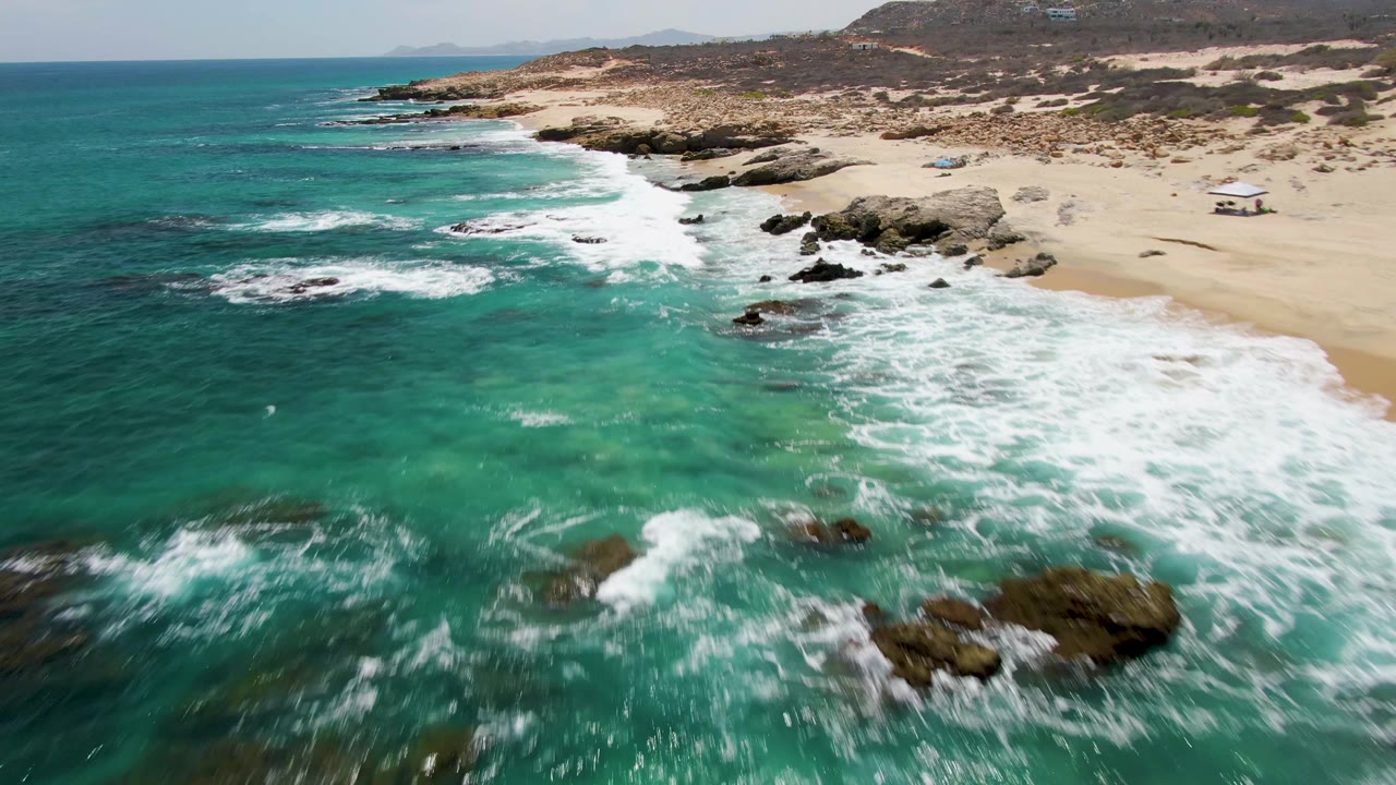 Aerial frame over Buzzards Beach in Baja California Sur, Mexico, showing white foam pushing toward the right edge, dark rocks in shallow green-blue water, sandy beach, low hills, and no visible people.
