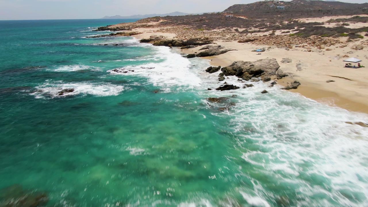 Aerial coastline view with foamy waves, rocks, sand, and turquoise shallows.