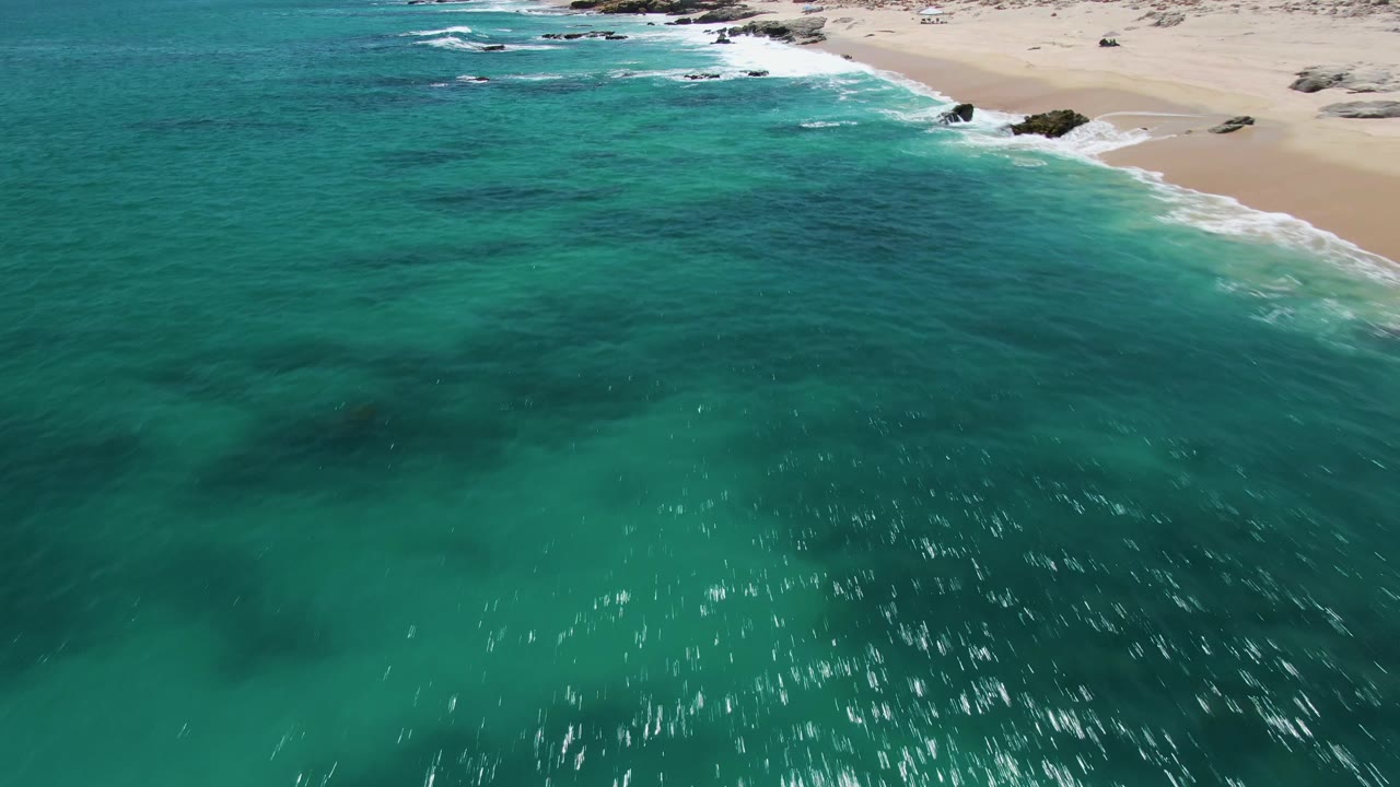 Aerial view over turquoise water at Buzzards Beach, Baja California Sur, with pale sand and scattered shoreline rocks at the top of frame and no visible people.