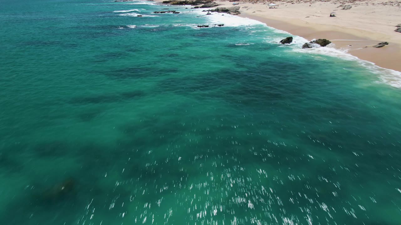 Aerial frame at Buzzards Beach showing rippled turquoise water, white foam along the sandy shore, dark underwater patches, and no visible people.
