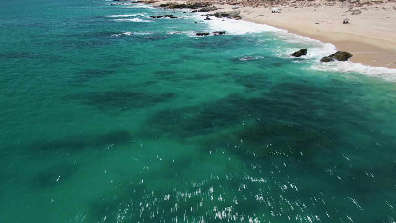 High oblique aerial of Buzzards Beach with black rocks interrupting the white shore break, turquoise shallows below, pale sand above, and no visible people.