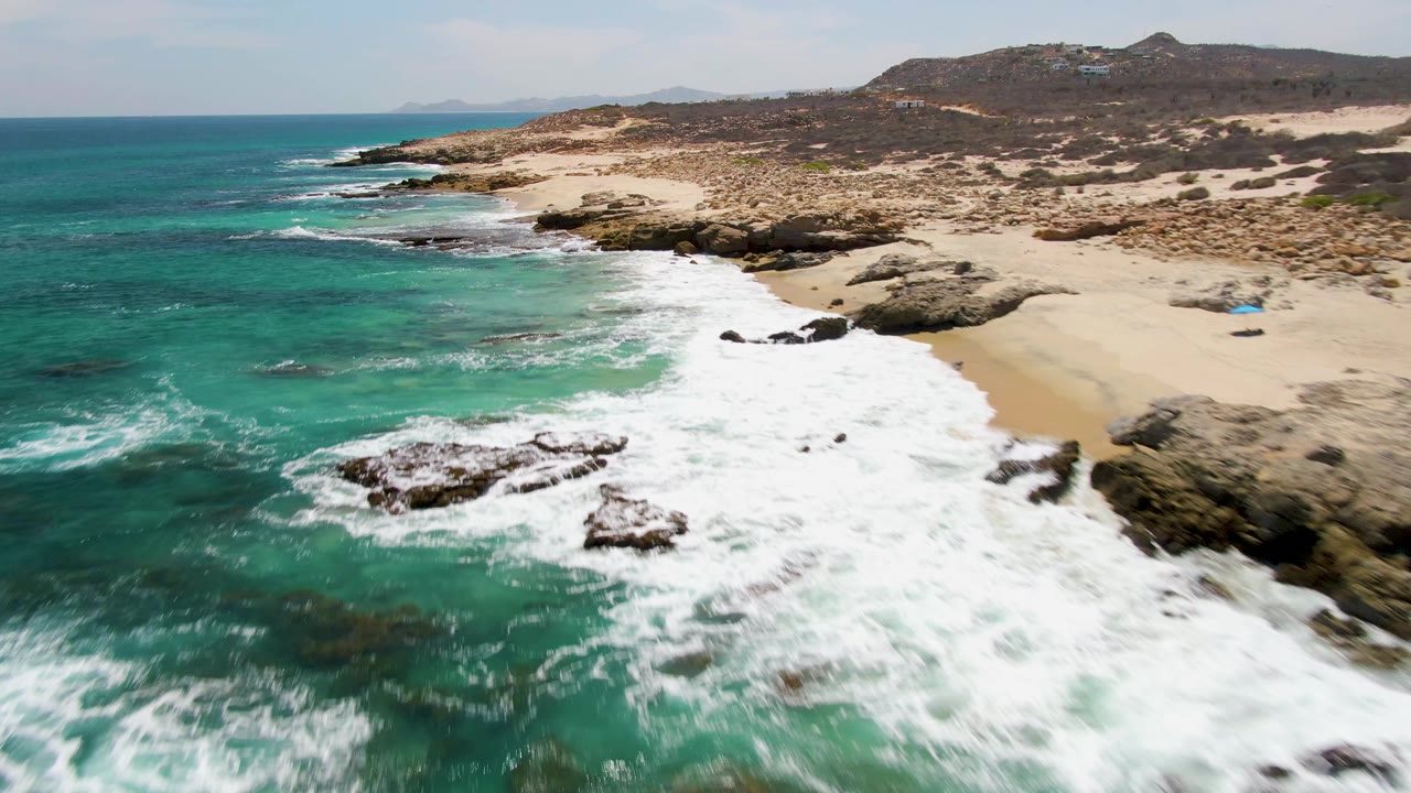 Aerial view at Buzzards Beach in Baja California Sur, Mexico, with turquoise water foaming over dark rocks, pale sand to the right, dry hills behind, and no visible people.