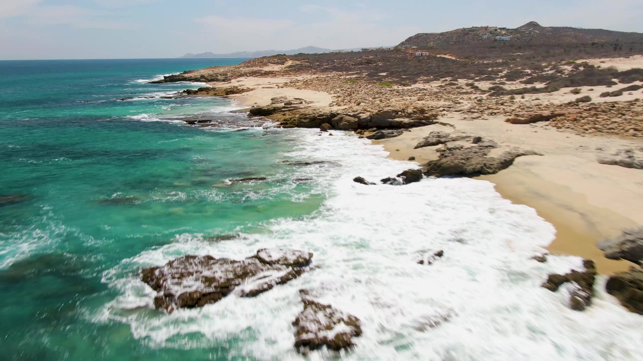 Aerial frame from Buzzards Beach showing a wide band of white surf over nearshore rocks, green-blue water on the left, sandy beach on the right, and no visible people.
