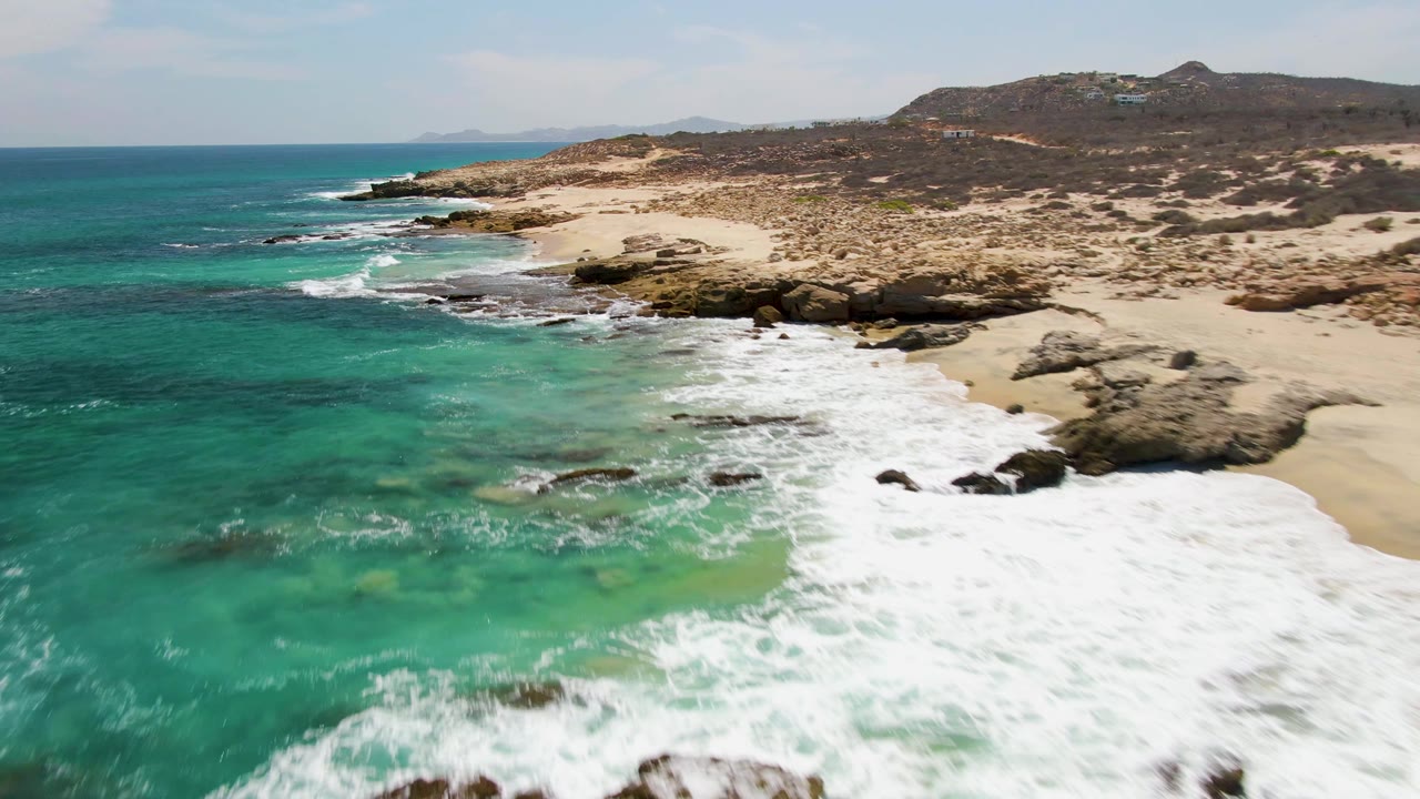 Aerial view at Buzzards Beach with more open turquoise water in the foreground, surf washing over rocks near the beach, low dry hills in back, and no visible people.