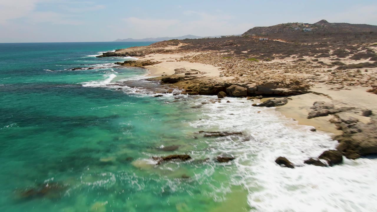 Aerial coastline with green water, breaking surf, rocks, sand, and scrub hills.